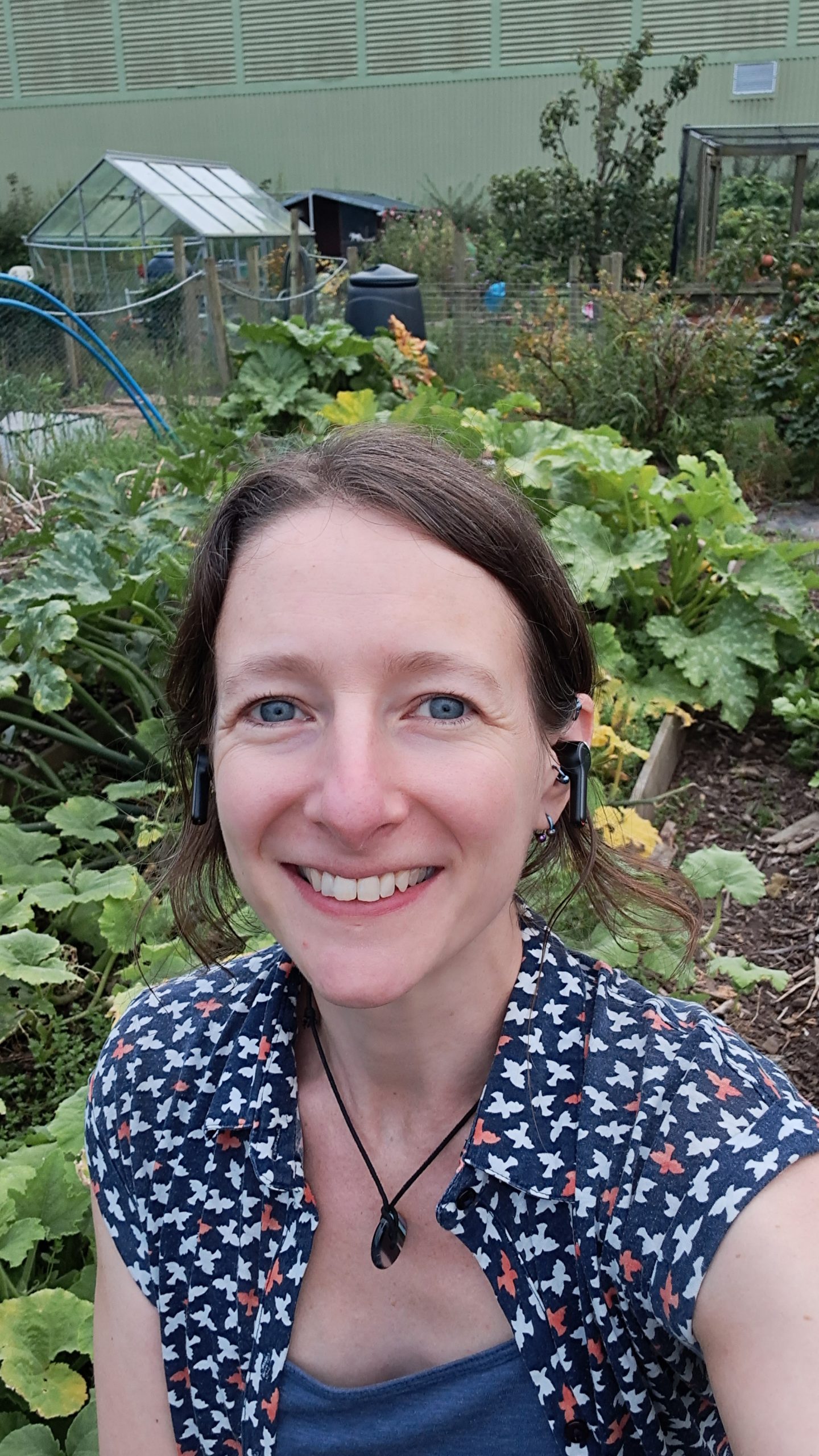 Becca, one of our tutors, smiling in front of a lush allotment garden with a greenhouse in the background.