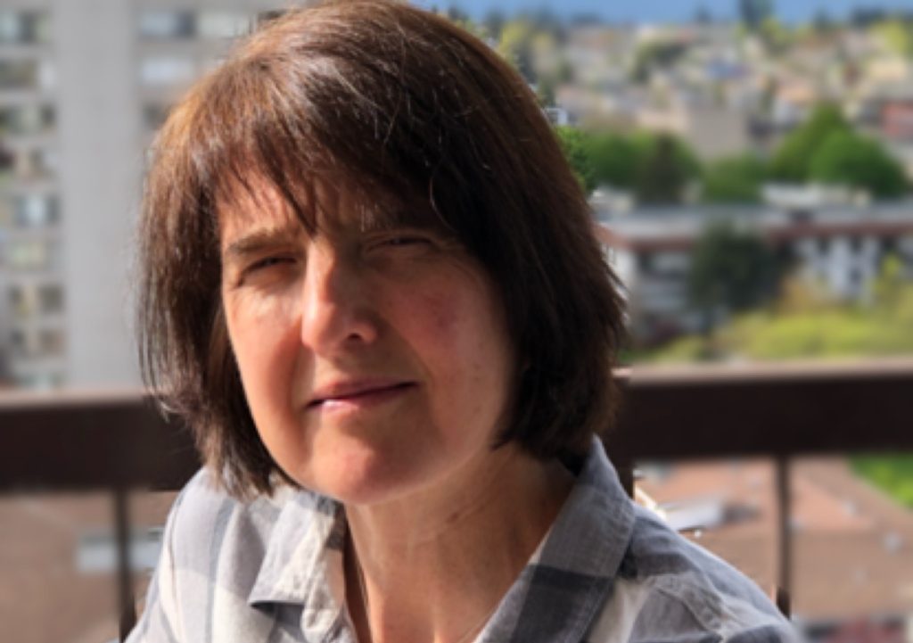 Tutor Mandy sits on a balcony wearing a grey checked shirt, with a blurred city and greenery in the background.