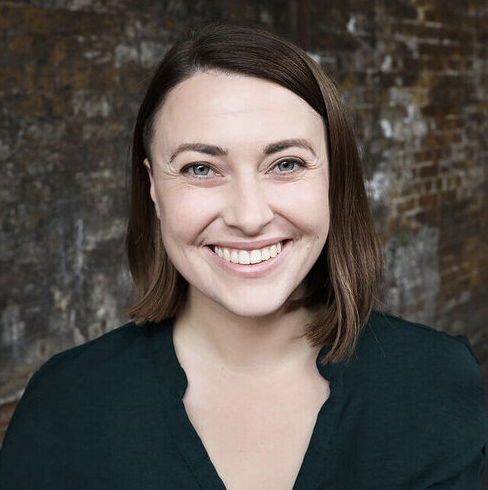 Tutor Rebecca smiling at the camera against a rustic brick wall, wearing a dark top.